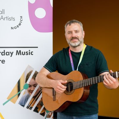 Guitar tutor in black t shirt holding guitar next to pop banner for GYA Norwich